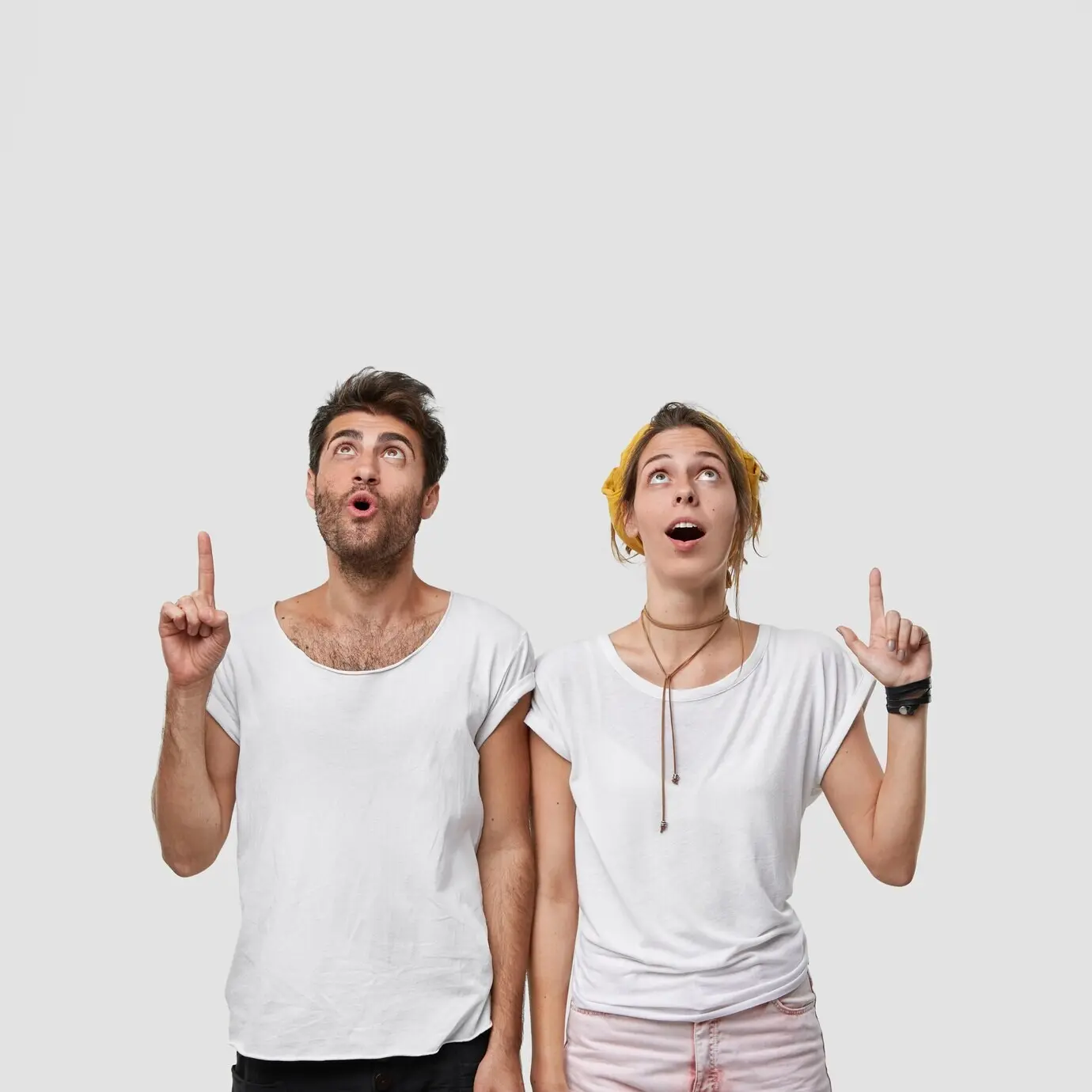 Vertical shot of a stupefied woman and man with jaws dropped, both pointing their index fingers upward to indicate something shocking, focused on the ceiling.